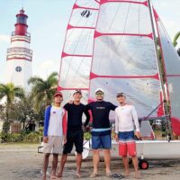 4 people posing in front of a Beneteau First 14 SE sailboat on a trailer in front of a lighthouse