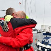 two men in red coats hugging in front of a Beneteau First 27 SE
