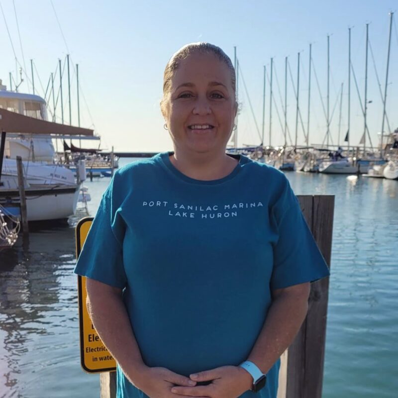 Tera Fox, smiling middle-aged woman wearing Port Sanilac Marina t-shirt in front of marina full of boats