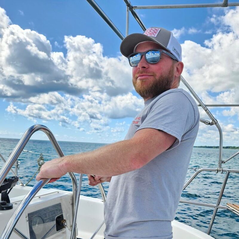 Jimmy, facilities manager. Middle aged man with a beard, hat and sunglasses helming a boat.