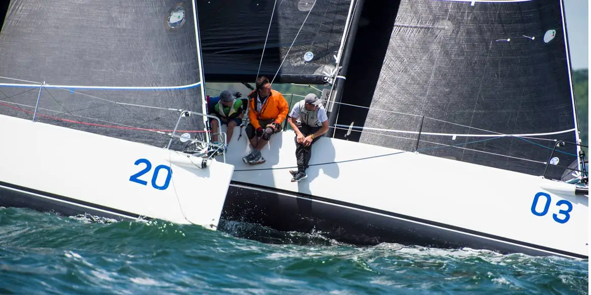 Three crew member leaning on side of a J-Boat J/111 sailboat right next to another boat during a race.