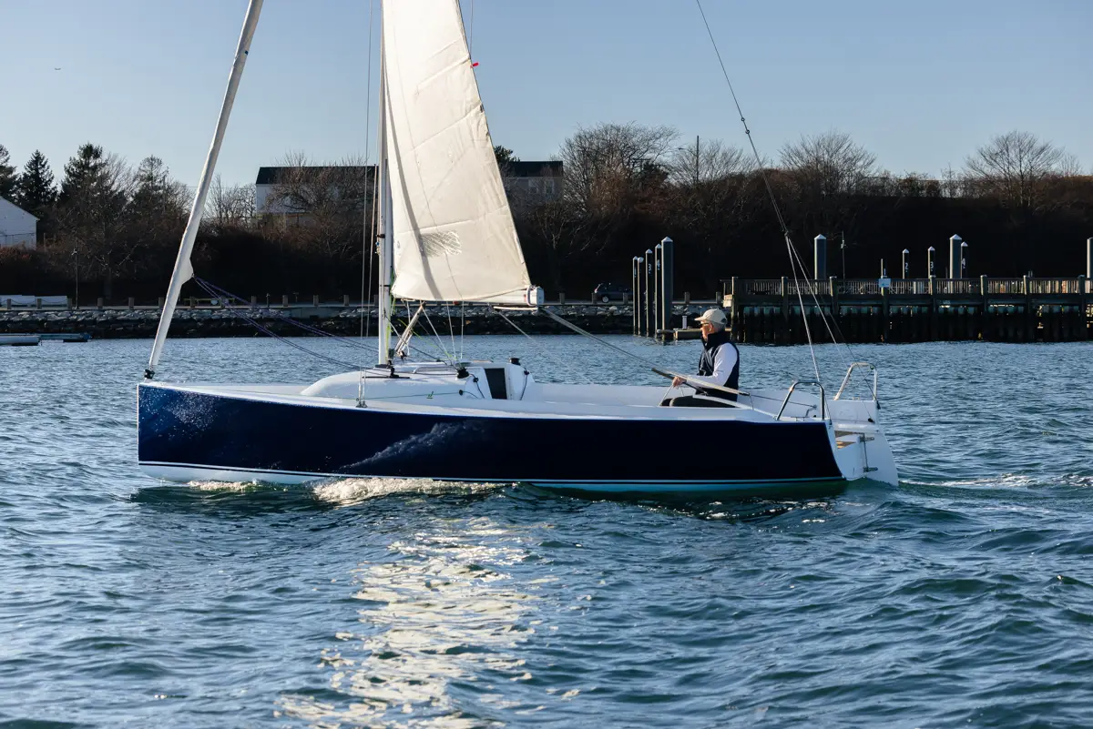 Older man sailing a J-Boat J-7 sailboat near docks.