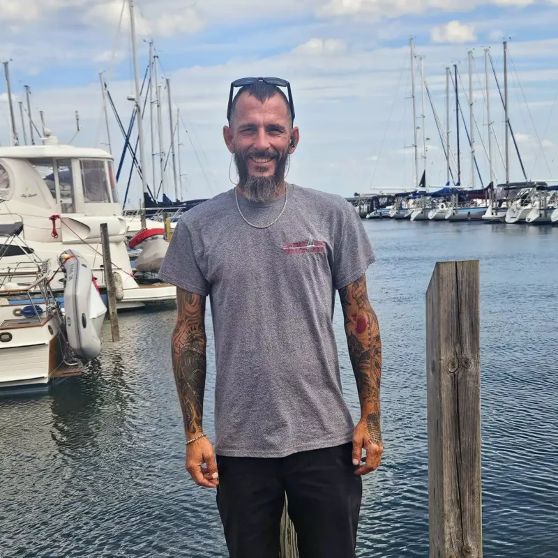 Service technician Dwayn, a smiling man with beard standing on a dock with sailboats docked behind him