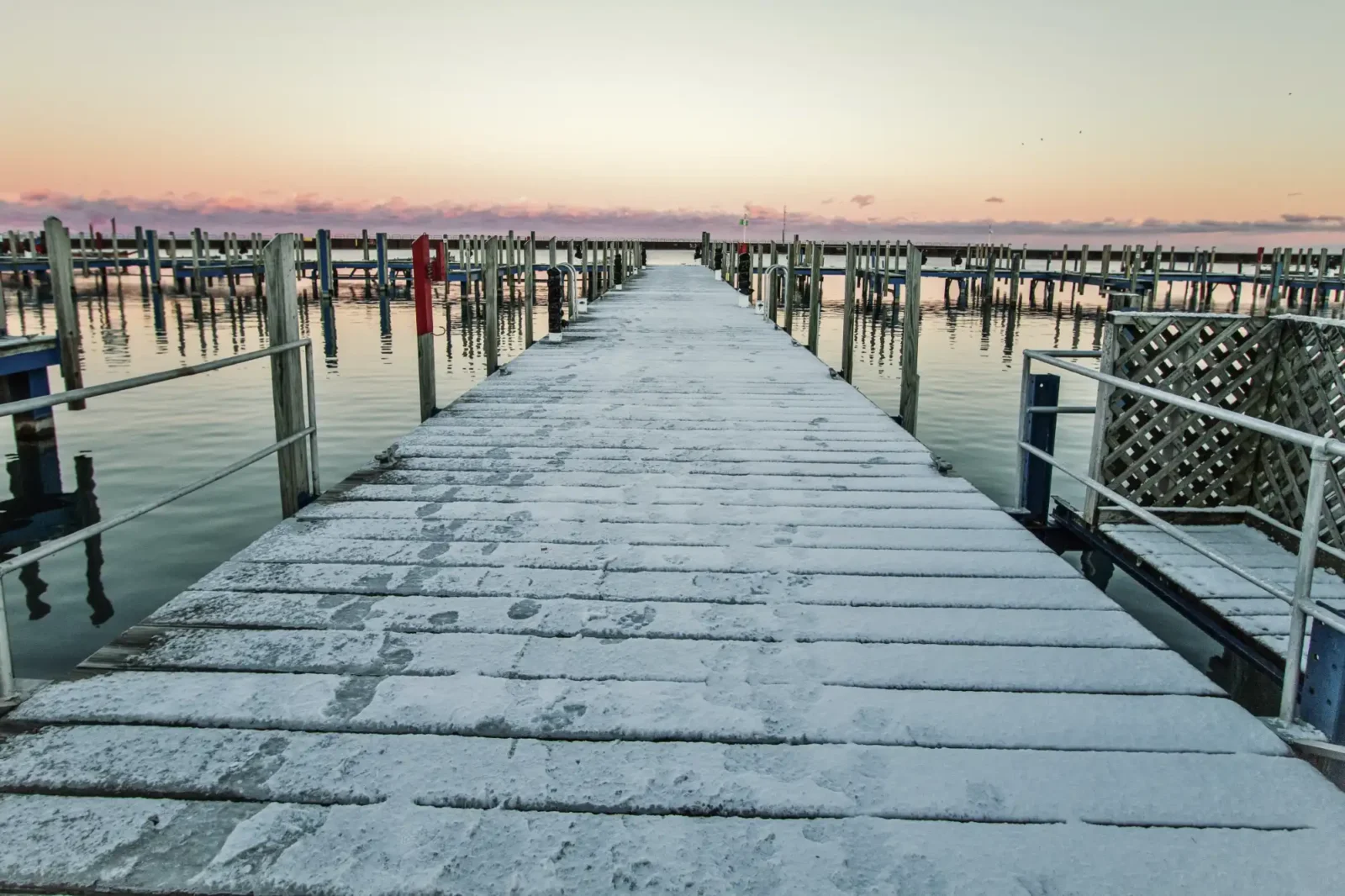 The snowy main dock at Port Sanilac Marina stretches into the distance, flanked by empty boat slips under a pastel winter sunrise.