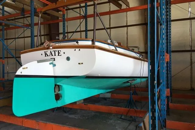 The stern of a clean 2007 Marshall 22 sailboat named KATE, with a white hull and teal bottom, is stored inside a marina building.