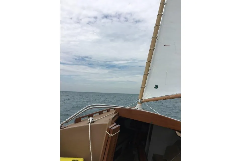 A low-angle view from the cockpit of a Marshall 22 sailboat underway, showing the white main sail and the ocean horizon beneath a cloudy sky.