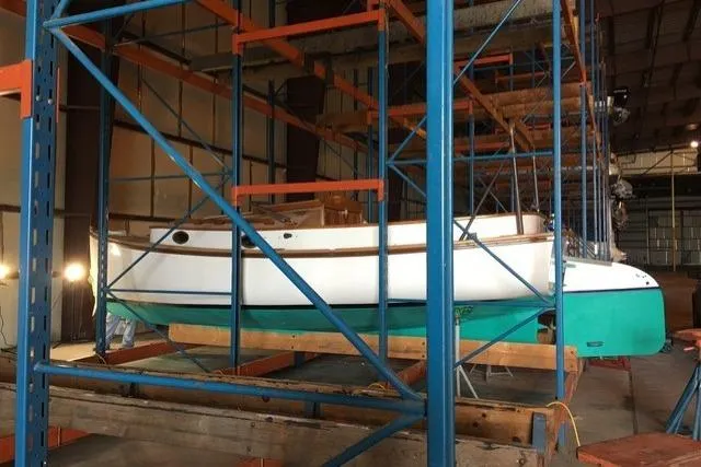 A white and teal Marshall 22 boat is elevated on wooden supports within the industrial racking system of a boat storage warehouse.