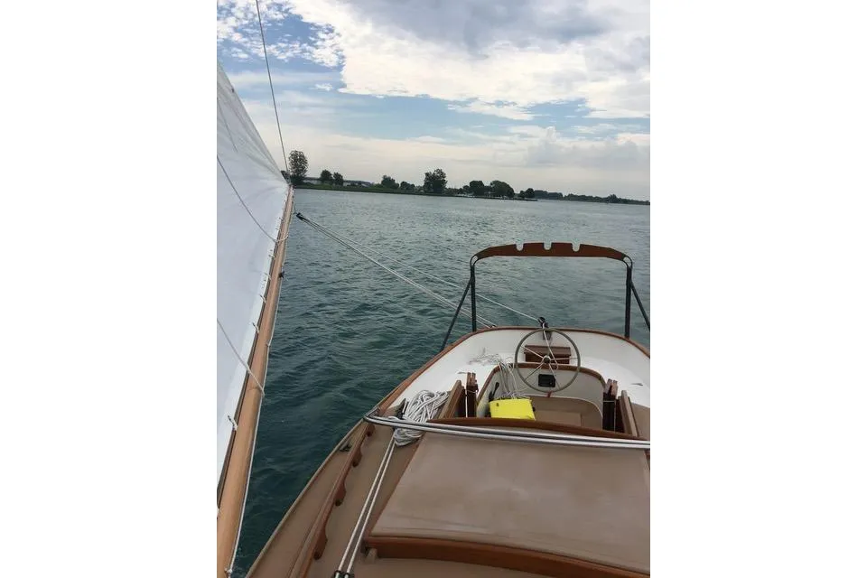 View from a wooden-trimmed sailboat deck, showing the helm and a white sail, sailing toward a tree-covered shoreline on choppy water.
