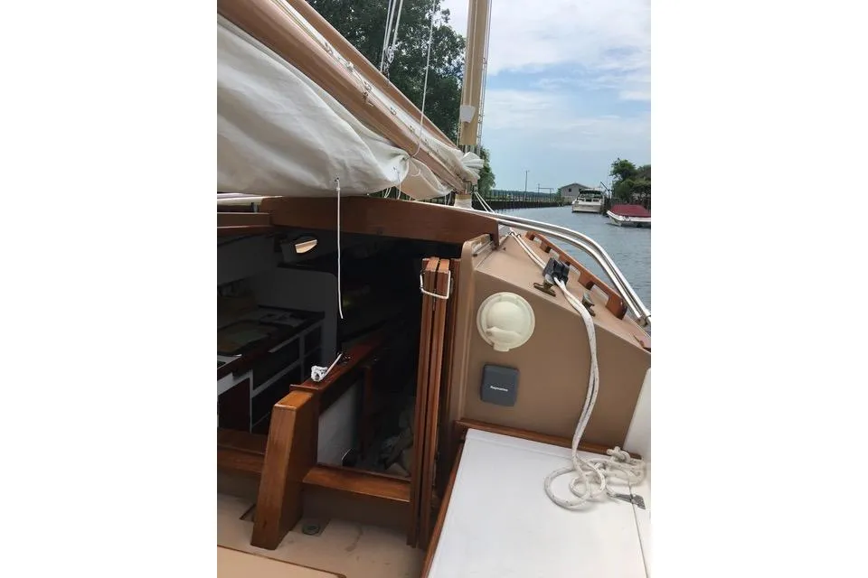The view from the cockpit of a catboat, highlighting the wooden cabin entrance, tan fiberglass, and the rolled white sail overhead while moored on a canal.