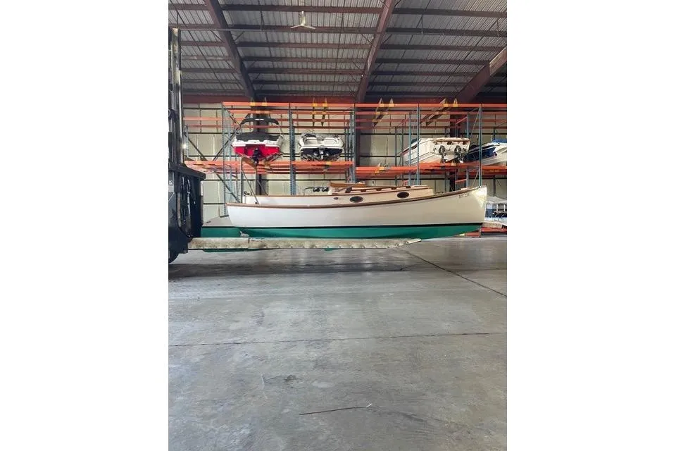 A white and wood-trimmed Marshall 22 sailboat with a teal hull is suspended by a forklift inside a large indoor storage facility. Other boats are stacked on racks in the background.