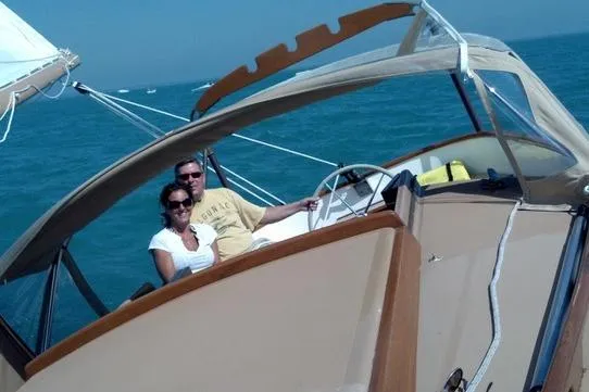 A smiling man and woman wearing sunglasses cruise together on a boat on the bright blue water, shaded by a light beige canopy.
