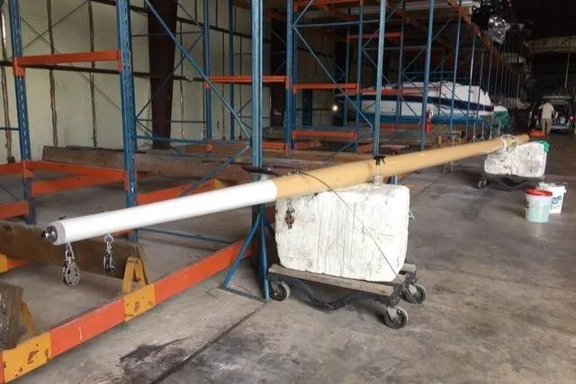 A lengthy boat mast, partially wrapped in white and brown material, rests on wheeled supports in a boat storage warehouse with high racks.