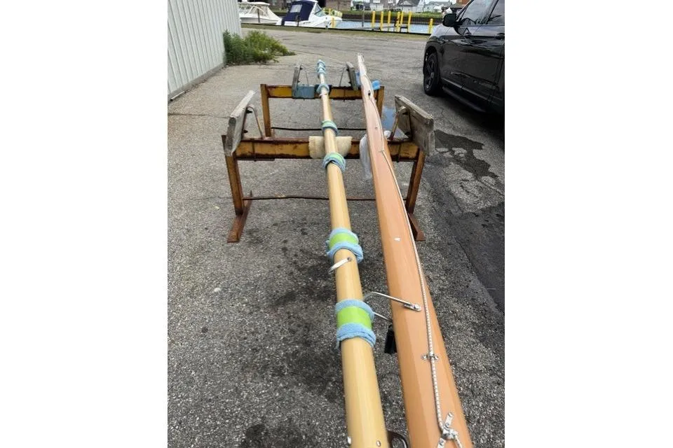 Two sailboat spars, a mast and boom, resting horizontally on a rusty yellow support stand in a marina yard. The mast is secured with blue and green padded wraps.