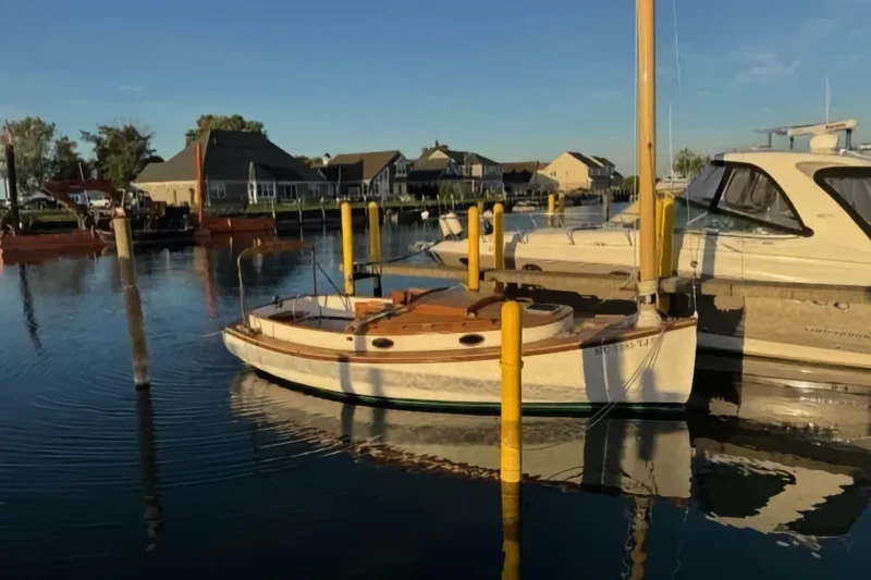 A white Marshall 22 sailboat with wooden trim is docked between yellow pilings in a marina, next to a yacht. Coastal homes overlook the tranquil water.