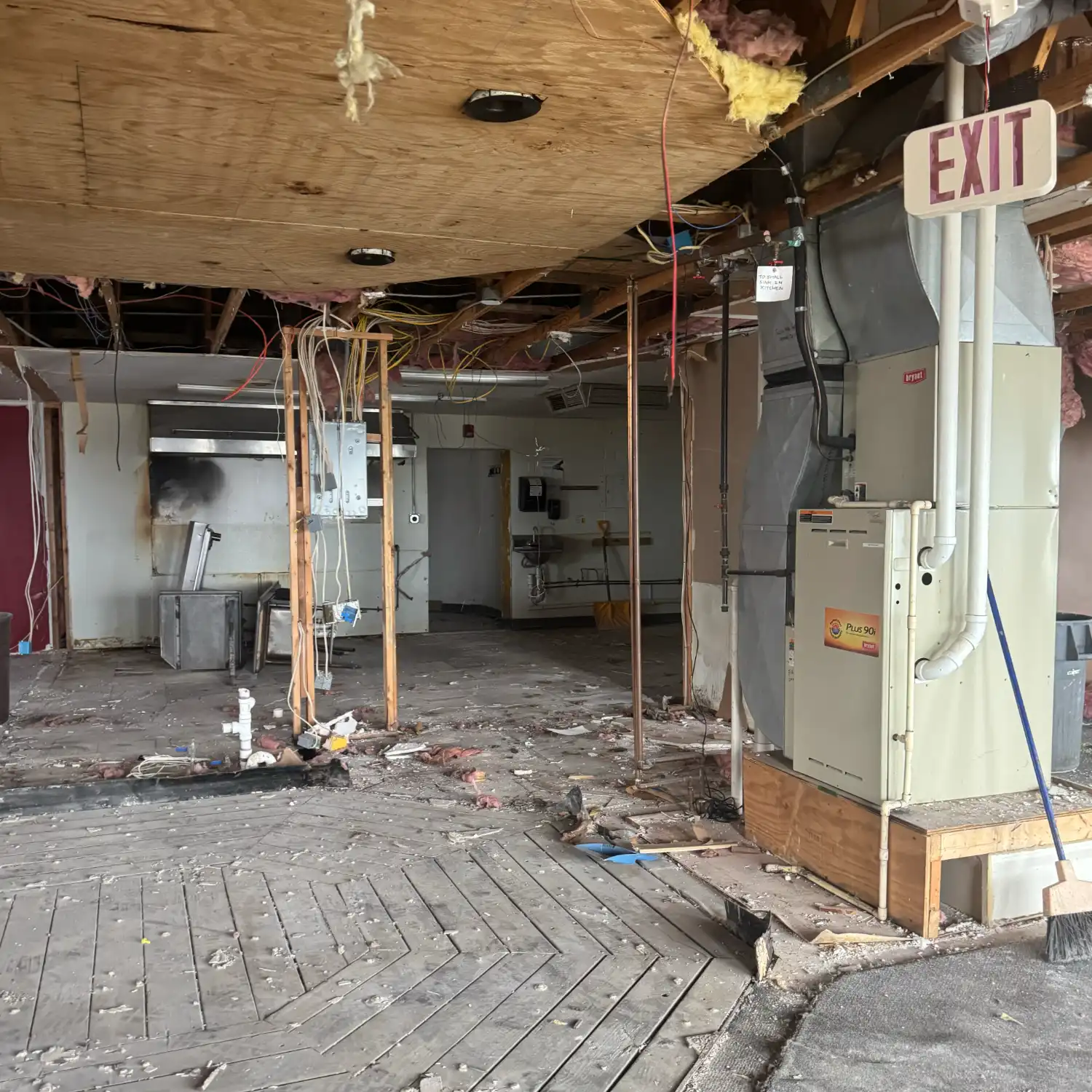 An actively renovating commercial interior showing exposed infrastructure, hanging electrical wires, debris on the wood floor, a large furnace unit, and an exit sign.