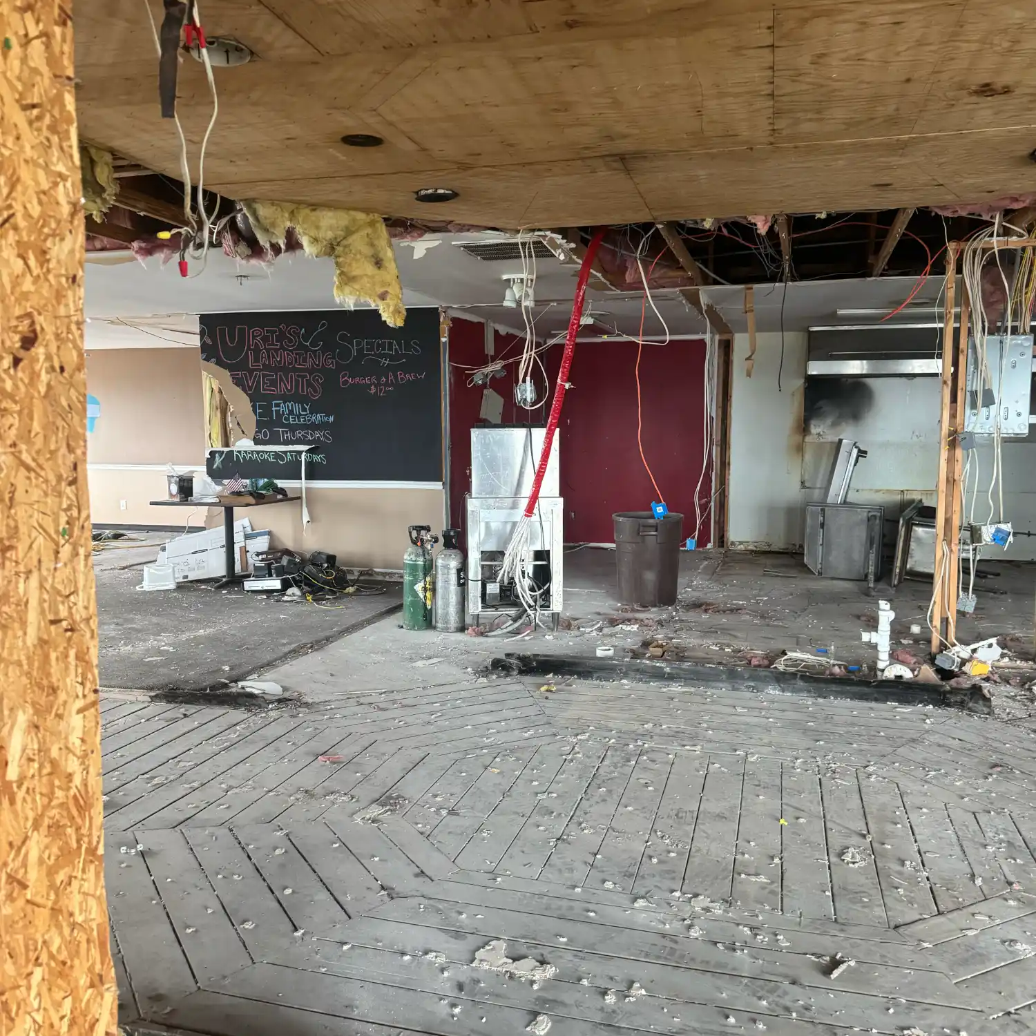 A commercial interior undergoing demolition, with an exposed ceiling, hanging wires, and wood subfloor. Dismantled kitchen equipment and a visible chalkboard menu are in the debris-filled room.