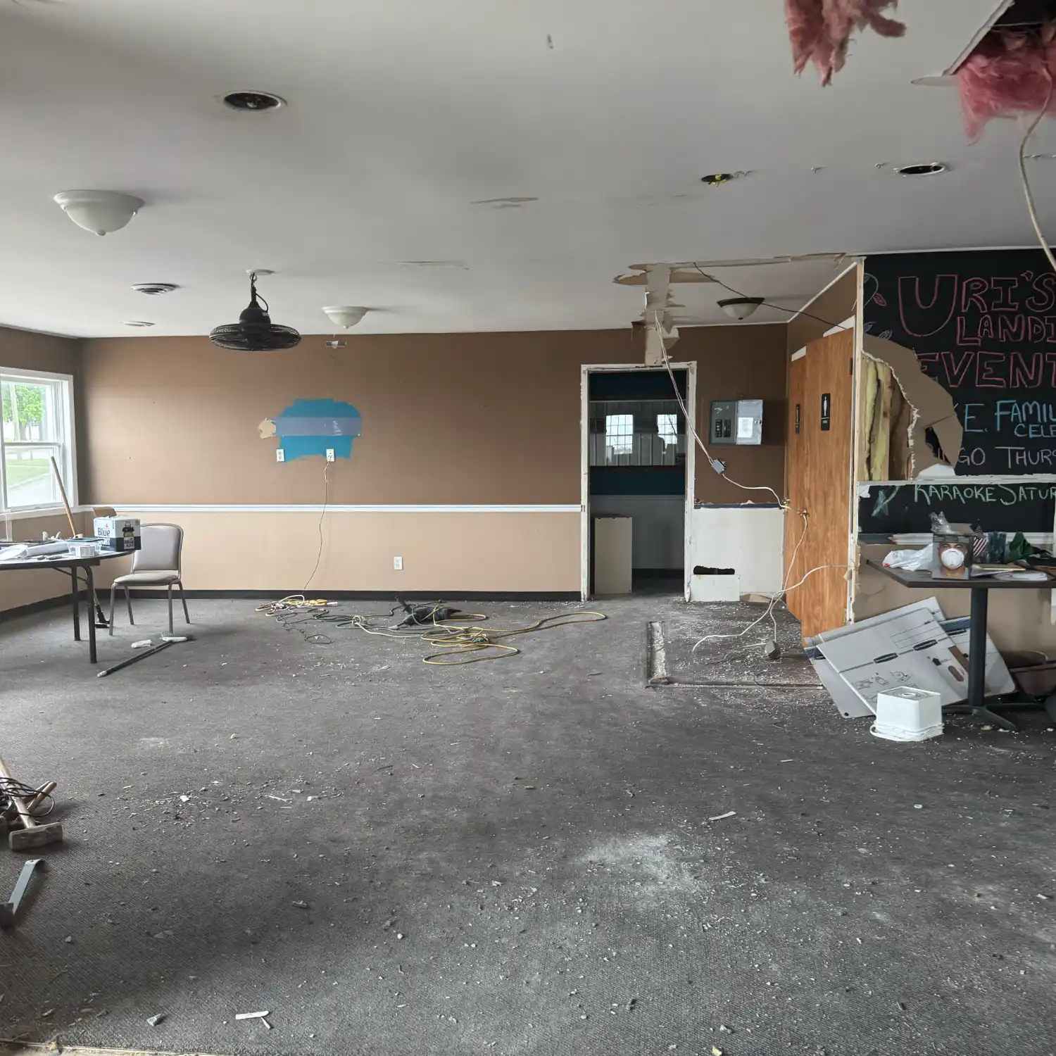 A commercial interior undergoing demolition, featuring debris scattered on the dark carpet, exposed wiring, and holes in the ceiling showing insulation.