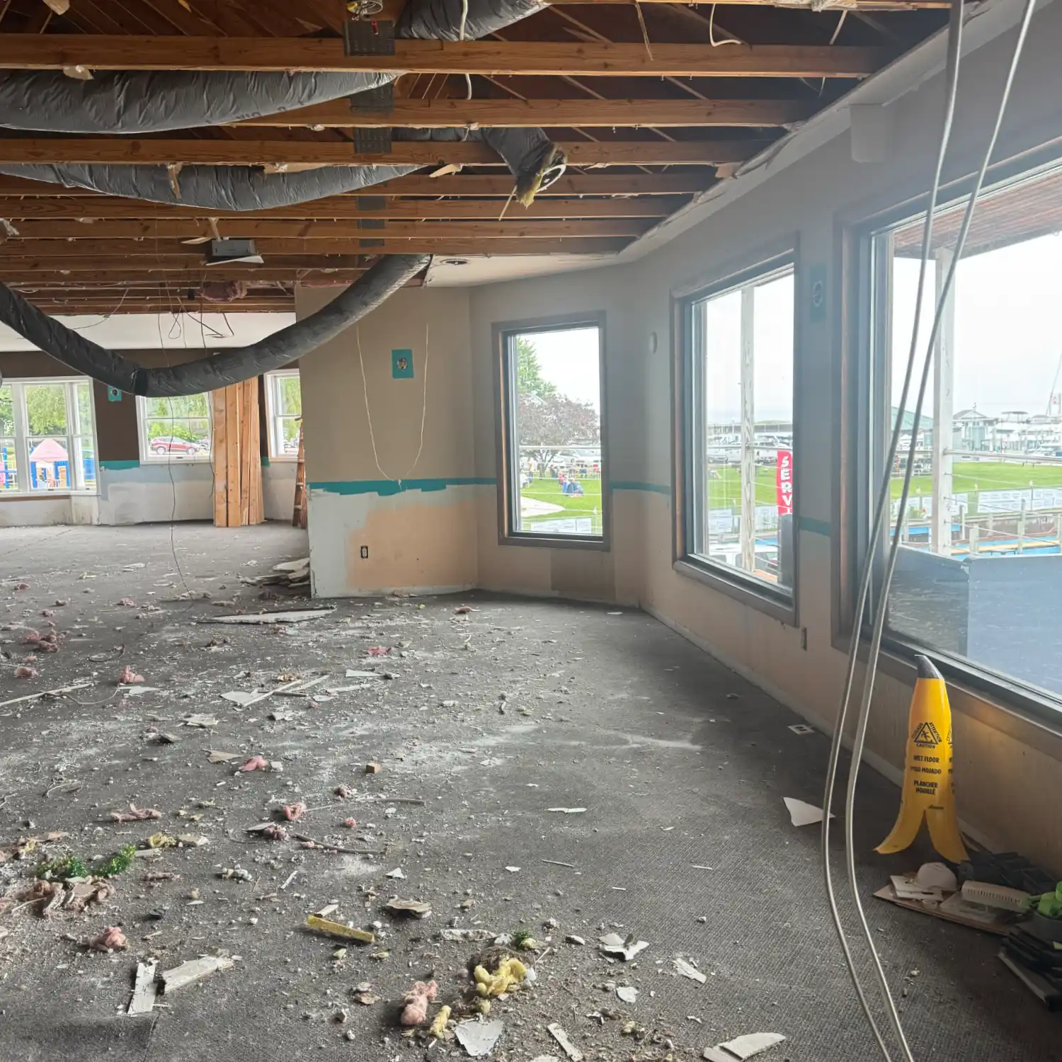 The interior of a commercial space during demolition, showing exposed ceiling beams, insulated ductwork, and debris covering the floor. Large windows overlook a marina.