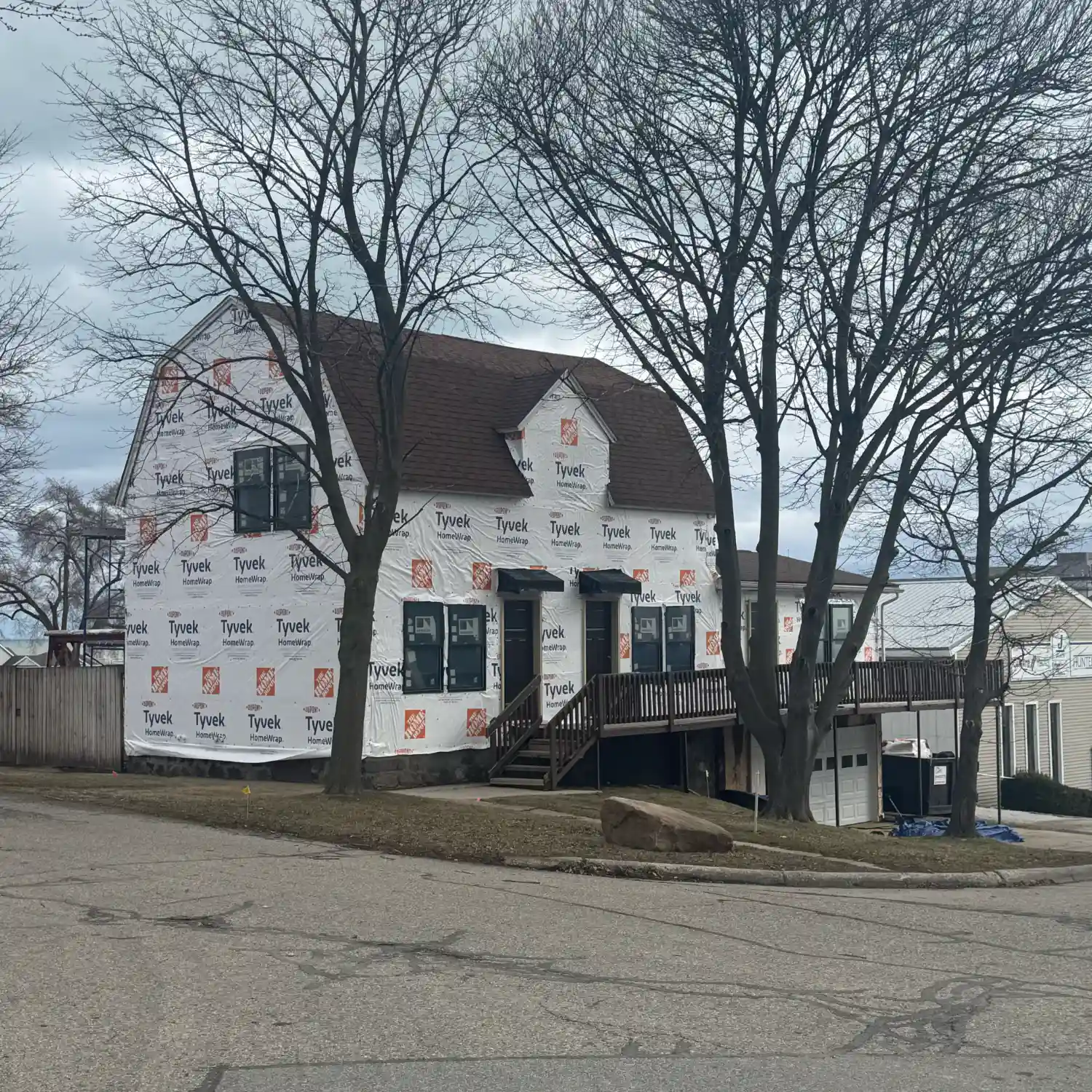 A large, two-story building undergoing renovation, completely covered in white Tyvek house wrap. It features a brown roof and an elevated wooden porch and stairway.
