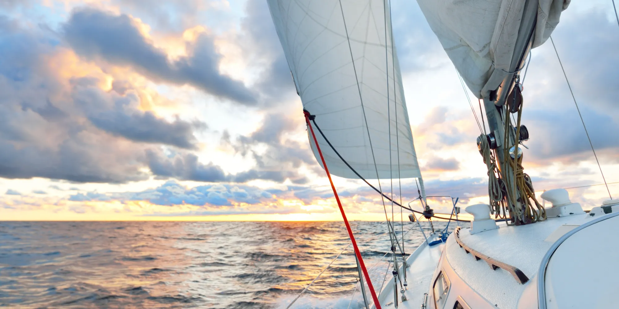 A close-up view from the deck of a white sailboat sailing into a dramatic cloudy sunrise. Warm orange light reflects on the taut main sail and choppy water.