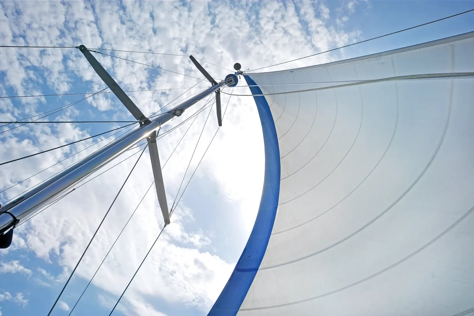 A view looking up the metallic mast and rigging of a sailboat. A white sail with a blue edge is visible against the bright, cloudy blue sky.