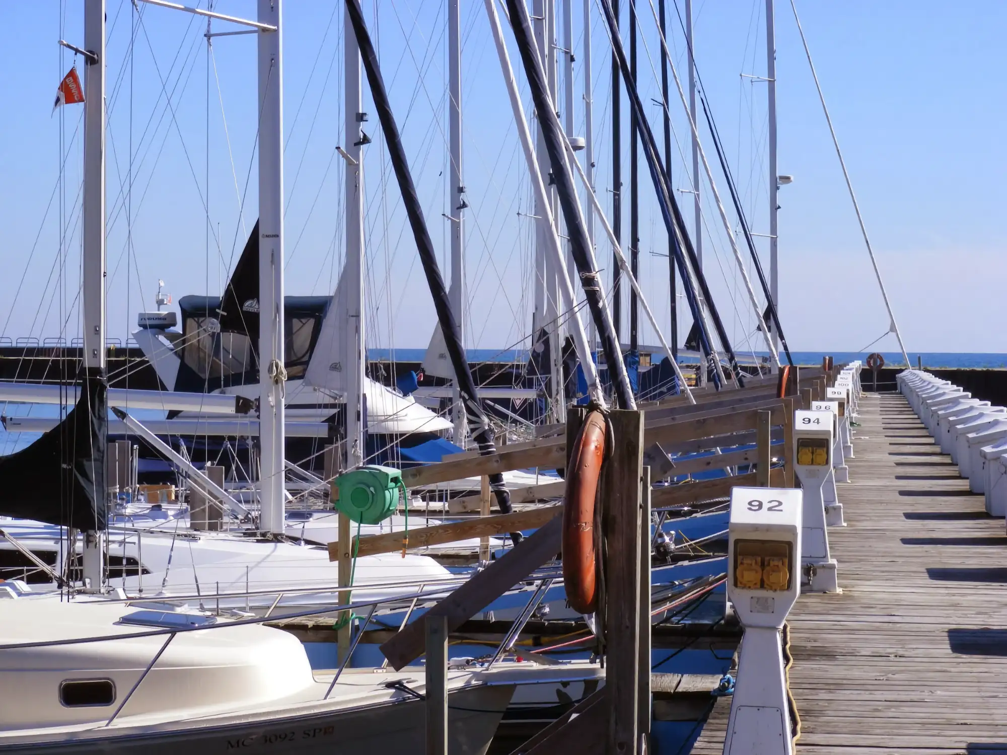 Sailboats are docked side-by-side in the Port Sanilac Marina. Tall masts reach toward the blue sky, viewed from a wooden dock lined with numbered utility posts.