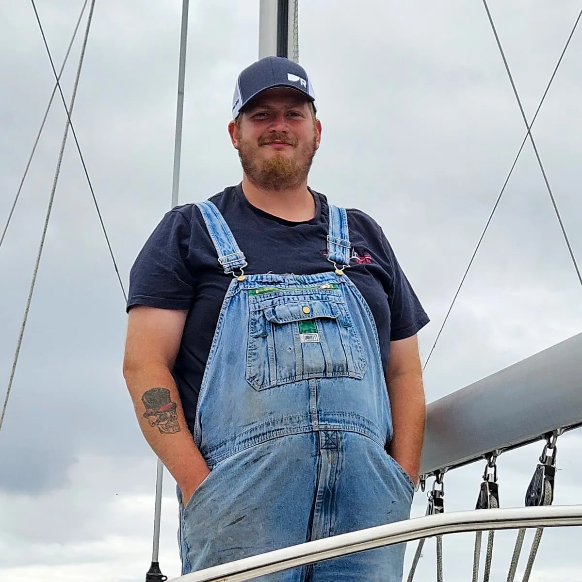 Service technician Austin Ebbs smiles outside, wearing denim overalls and a baseball cap near nautical rigging.
