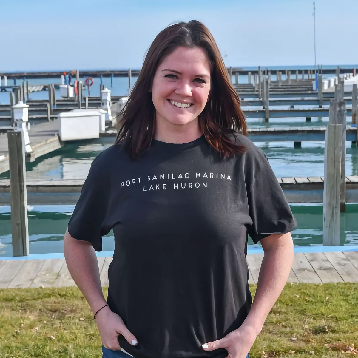 Jillian Sheldon, General Manager of Port Sanilac Marina, stands smiling in a dark t-shirt in front of the docks overlooking Lake Huron.