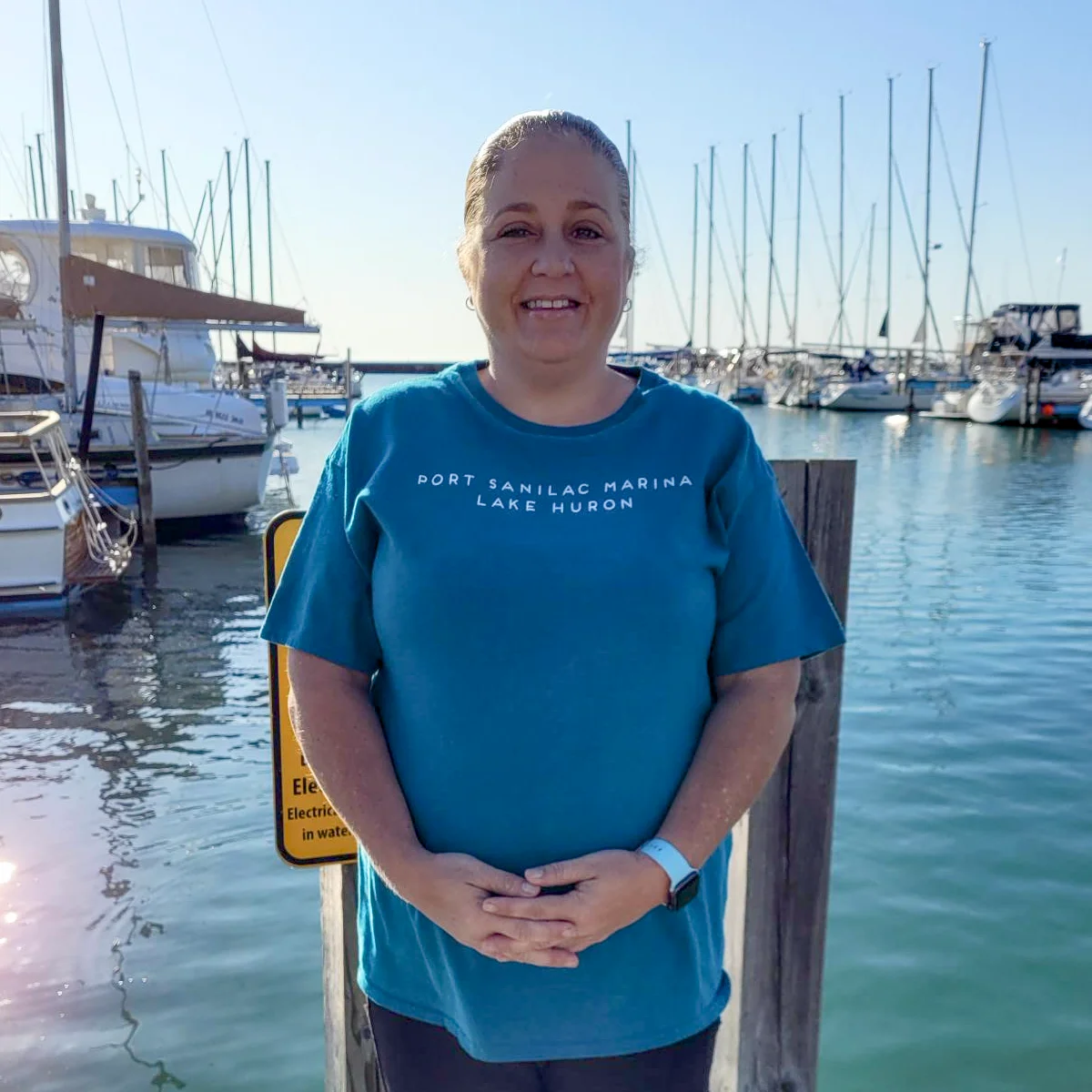 Tera Fox smiles on the dock at Port Sanilac Marina. She is wearing a teal t-shirt, standing in front of several moored sailboats on a sunny day.