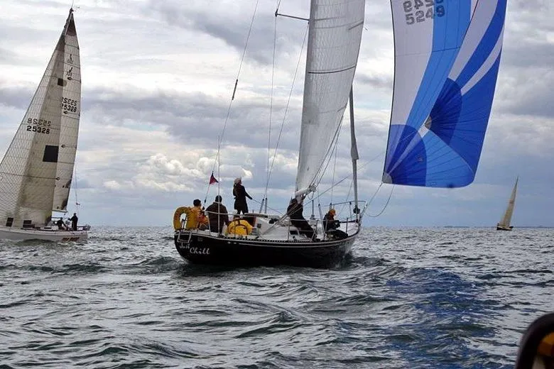 A black sailboat, crewed by six people in foul-weather gear, sails on choppy waves with a blue and white spinnaker deployed during a race under a cloudy sky.