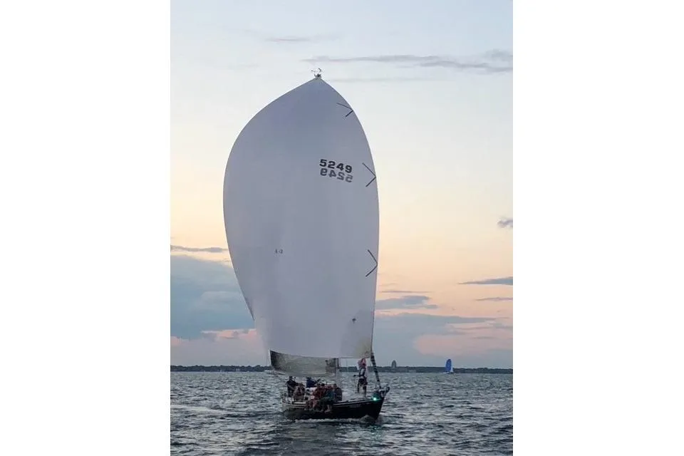 A sailboat marked 5249 with a large white sail sails on the water at twilight. A crew is visible on the deck against the soft, colored sky.