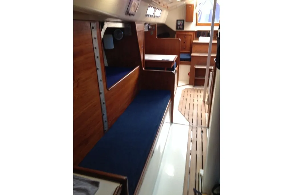 A clean boat cabin with rich wooden trim, dark blue bunks along the port side, a small table, and a wooden grate mat near the companionway.