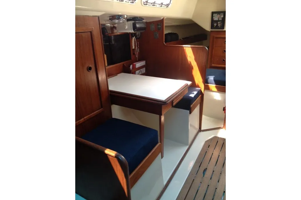 The interior of a sailboat cabin showing a polished wood dinette with a white table and dark blue cushions. The warm wood paneling is lit by bright sunlight.