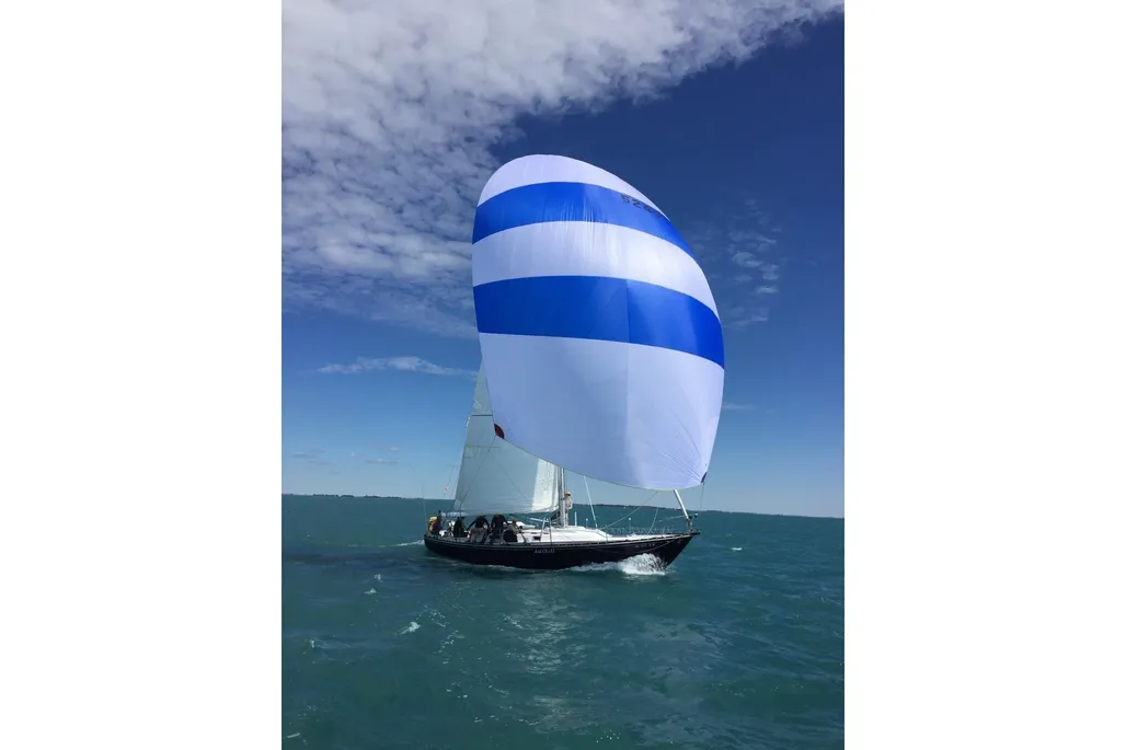 A dark sailboat speeds across the water, powered by a large white spinnaker sail with bold blue horizontal stripes against a bright blue sky.
