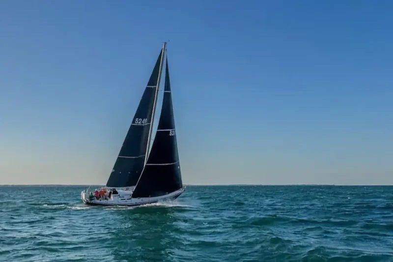 A white sailboat featuring large black sails races across choppy blue ocean water under a bright, clear blue sky.