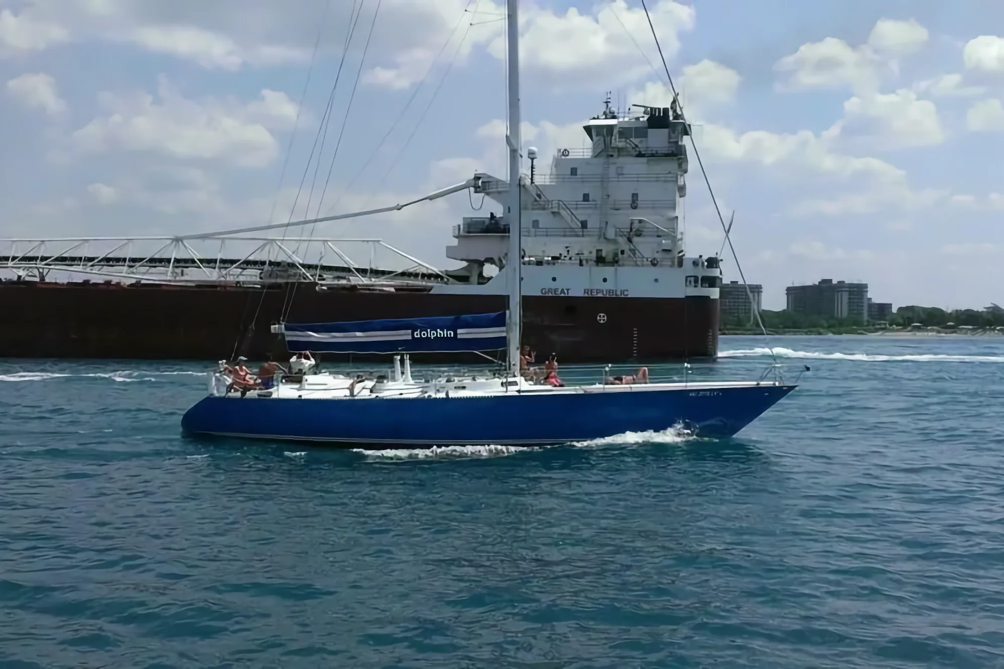 A blue sailboat named Dolphin, carrying people relaxing on deck, sails across the water past the massive Great Republic cargo ship on a clear day.