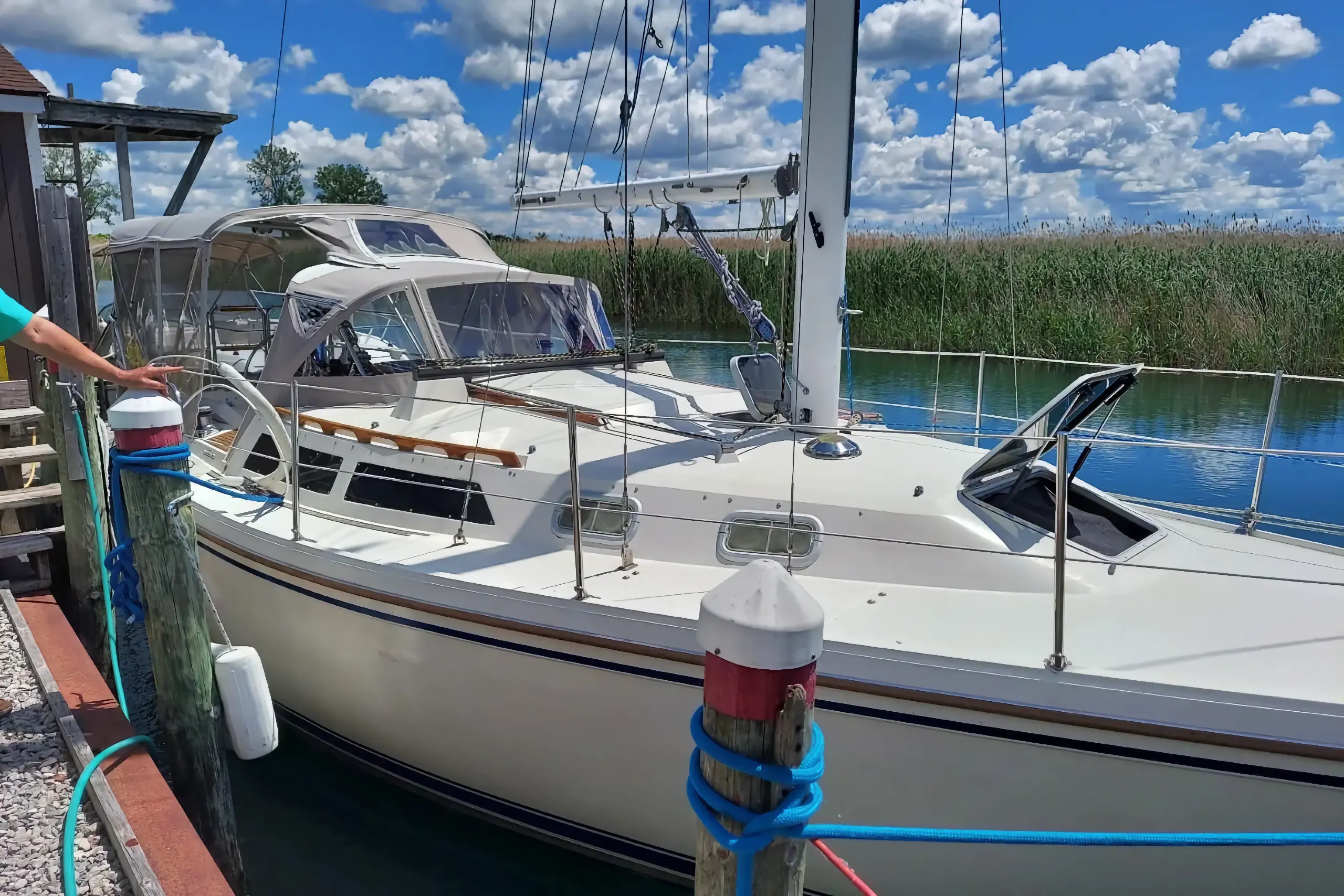 A white sailboat with a gray cockpit cover is securely docked to a wooden piling. The marina channel features calm water and tall green reeds under a blue, cloudy sky.