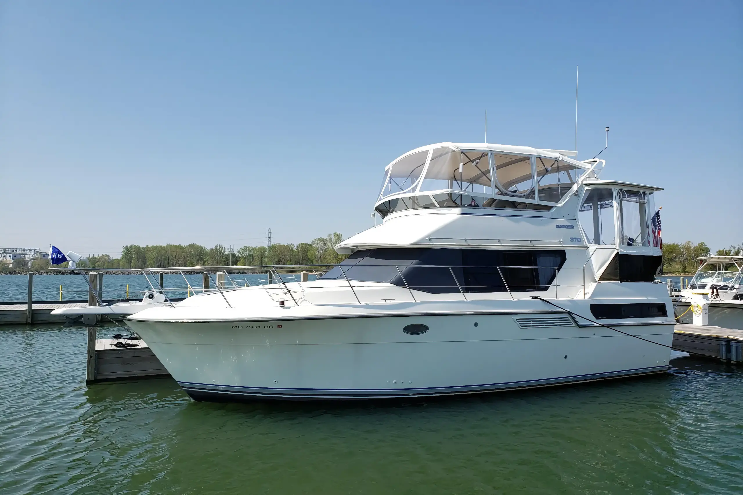 A large white Carver 370 Aft Cabin Motor Yacht with a clear flybridge enclosure is docked in a sunny marina, secured between wooden piers on green water.