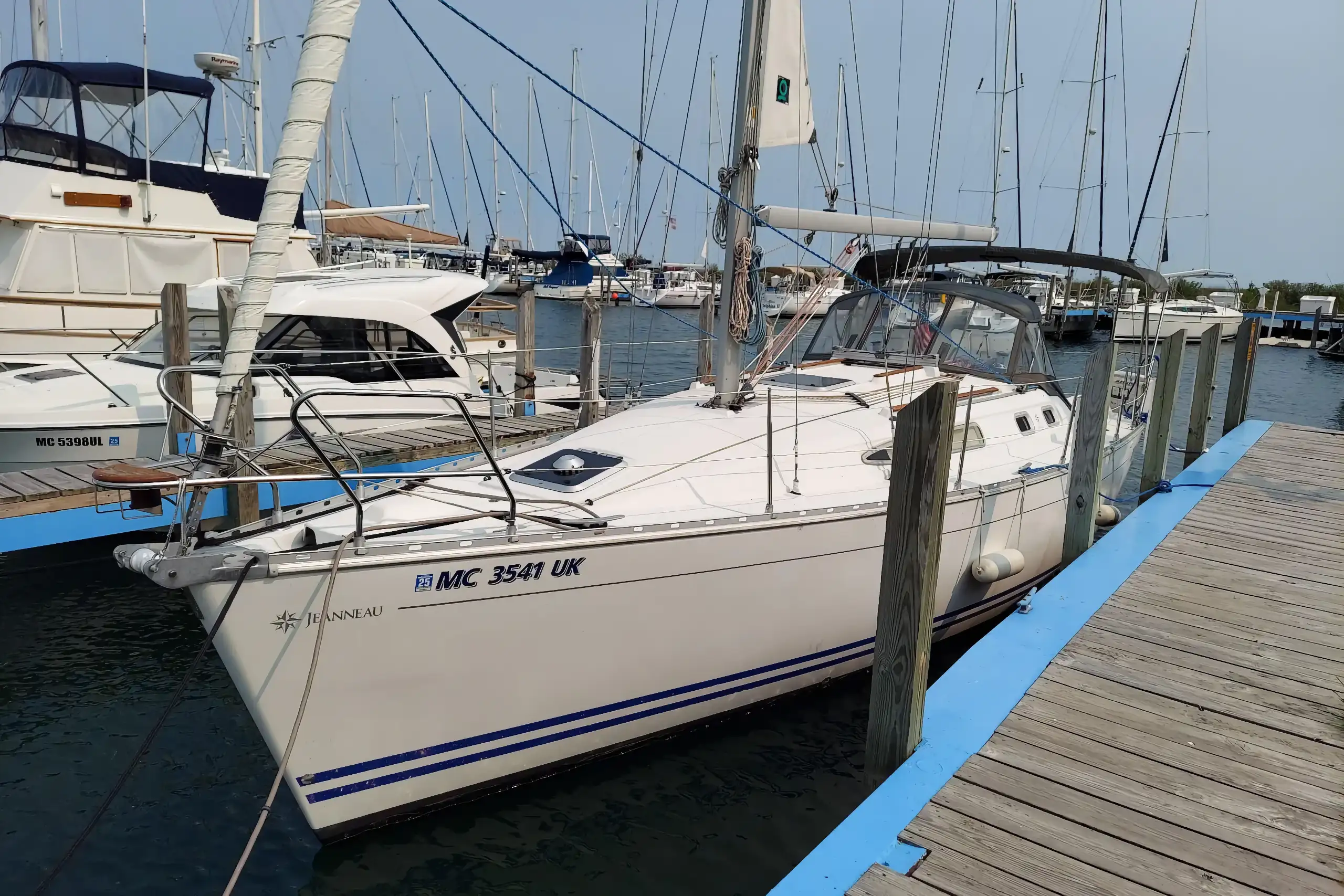 A white 2000 Jeanneau 34.2 sailboat is securely docked along a wooden pier in a busy marina under a bright sky, surrounded by other masts.