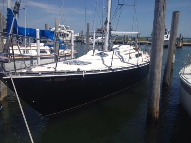 A dark-hulled sailboat with a white deck and rigging is securely moored in a marina slip between large wooden pilings. Other boats are visible in the background.