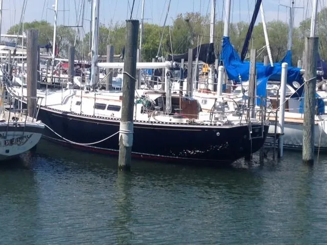 A view of a sailboat with a dark hull and white deck docked at a marina among other boats. Wooden pilings secure the boat in the calm water.