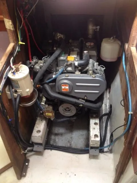 A gray marine engine is mounted on supports inside a tight wooden engine compartment, surrounded by hoses, electrical wiring, and a white expansion tank.