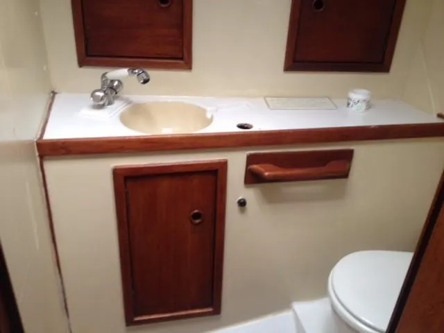 A compact marine head interior showing a beige wall and dark wood trim. It features a white vanity, a light sink basin, and a partial view of the toilet.