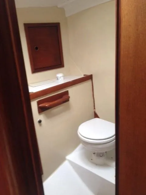 The small, clean marine head of a boat, visible through a wooden doorway, featuring a white toilet, cream walls, and dark wooden cabinets and trim.