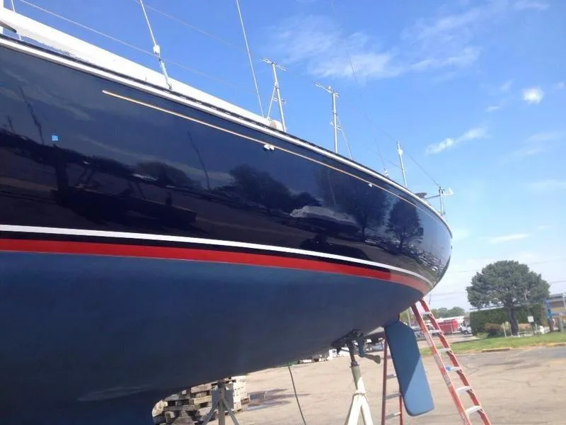 The side of a large sailboat hull, polished to a high-gloss navy blue, sits on stands against a bright blue sky. Red and white stripes mark the waterline.