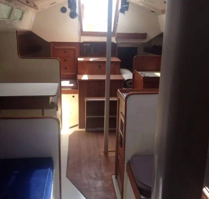 The bright interior of a boat cabin features dark wood steps leading forward, varnished wood floors, white bulkheads, and seating areas with blue cushions.