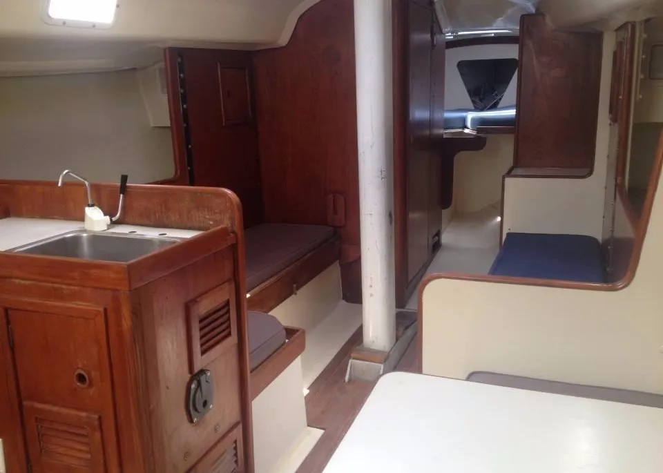 The main saloon of a boat cabin, featuring rich wood cabinetry and paneling, a small galley sink, and settees extending towards the forward V-berth area.