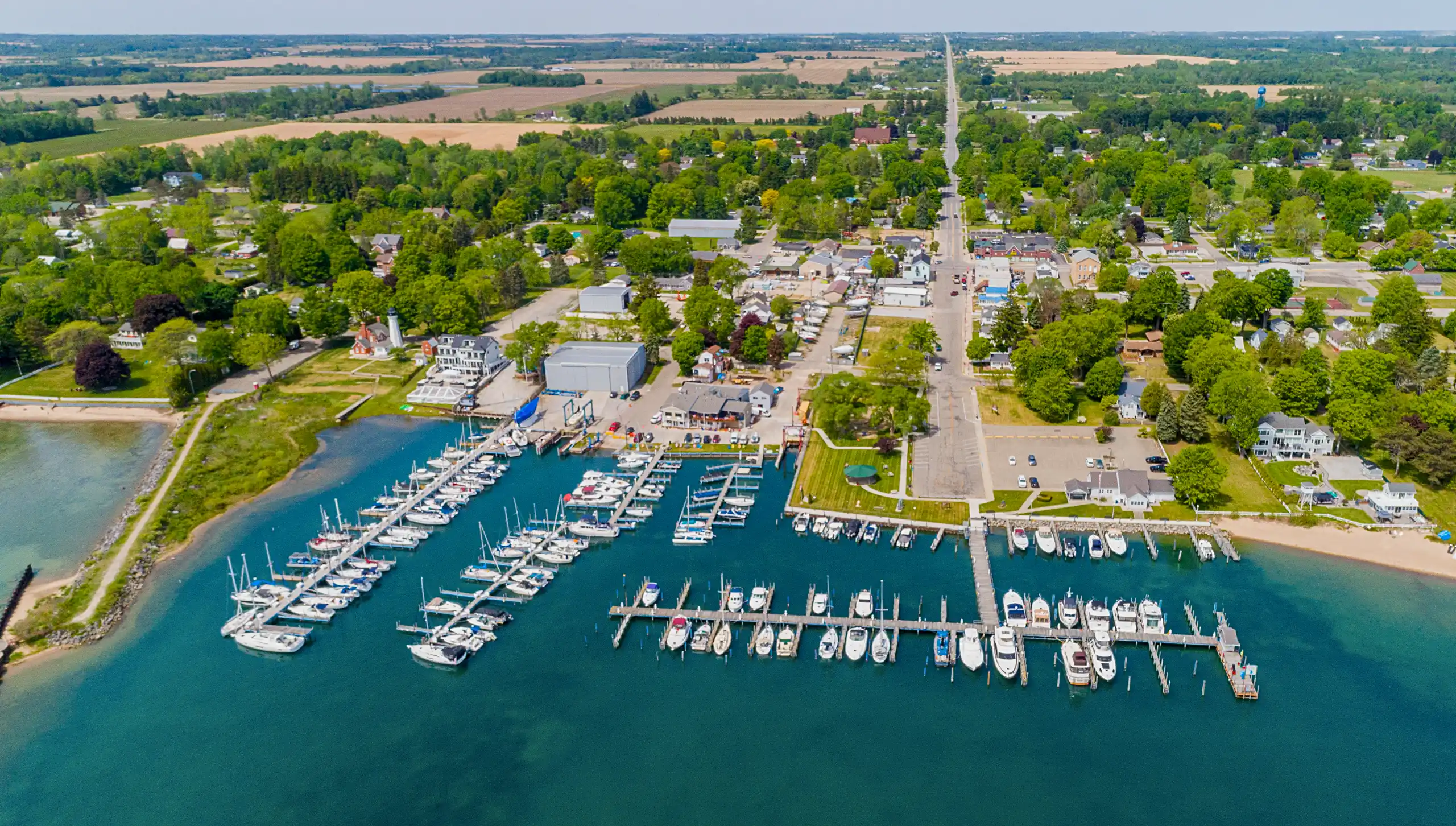 An aerial view of a busy marina filled with docked boats in turquoise water. The waterfront town extends inland along a main road surrounded by green trees and agricultural fields.