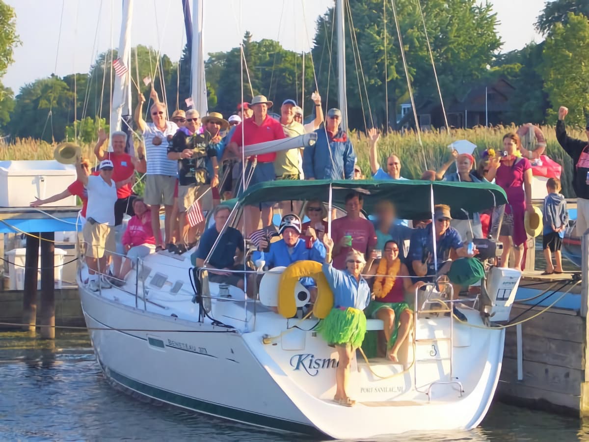 A lively crew, some wearing hula costumes, celebrate and wave their hands and small American flags aboard a docked sailboat.