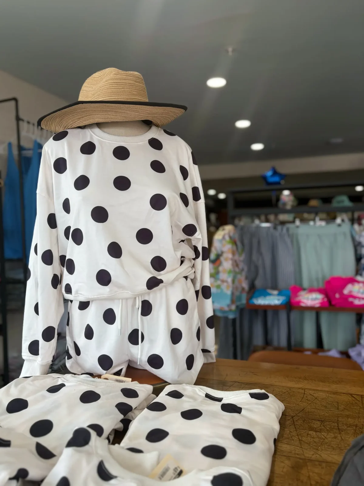A mannequin wears a white, black polka-dot loungewear set (long sleeve top and shorts) and a straw hat with a black brim in a store display. Folded matching clothes are on the table.